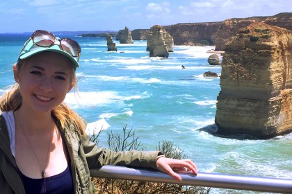 Girl with blond hair and hat on rest arm on railing in front of a backdrop of blue ocean and rocks.