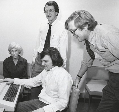 Black and white image of four people gathered around a computer