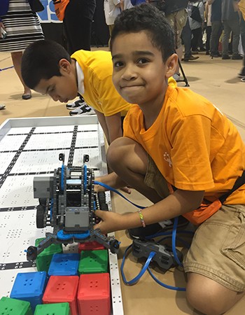 A boy in a yellow shirt holds a small robot with gears and metal parts, which scoops colored plastic blocks