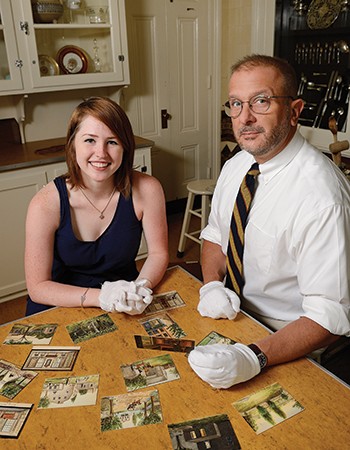 A student and a man, both wearing gloves, sit at a table covered with old miniature paintings