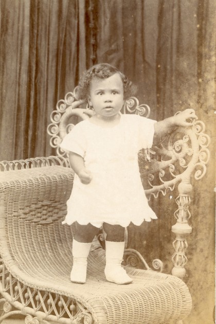 Sepia photo of small child standing on a chair. She wears a dress and boots.