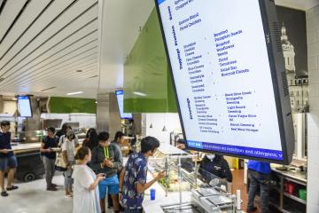 A digital screen displays a menu while students order food in the background