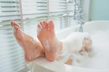 Woman’s feet rest on the top edge of a bathtub. Her legs are surrounded by bubbles.