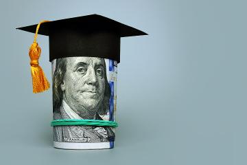 A graduation cap with tassel sits atop a roll of cash showing Benjamin Franklin’s face