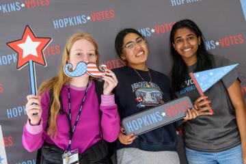 Three students pose for a photo, one holding a star and U.S.-flag-themed mustache prop, another with a Go Vote pennant, the third with a Hopkins Votes sign