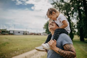 A heavily tatooed man is looking up and smiling at his young daughter sitting on his shoulders.
