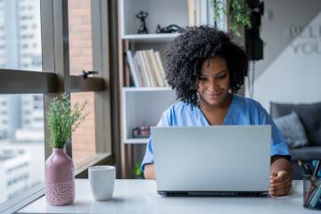 A nurse in scrubs uses a laptop computer in a home office