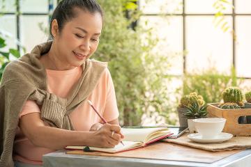 Middle-aged woman sitting at a table and writing in a journal.a coffee cup is on the table next to her.