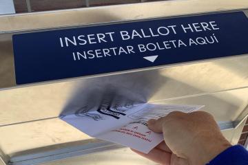 A hand inserts a ballot into a ballot drop box