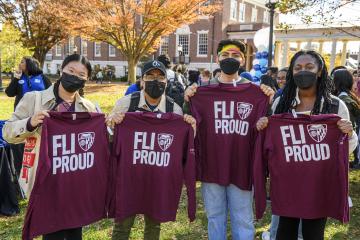 Four students stand side by side hold maroon FLI Proud t-shirts