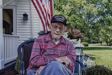 A man wearing a veteran's cap sits with a flag in the background and a walker in the foreground