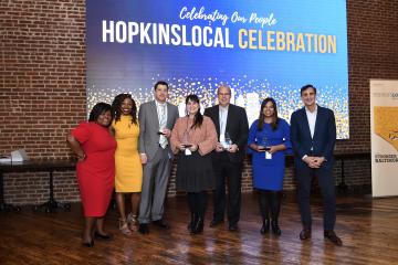 A group of HopkinsLocal award winners stands before a blue backdrop in front of a red brick wall