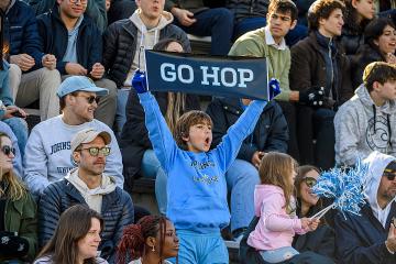 A group of Hopkins fans in the stands at a recent lacrosse game. A young boy in the front is dressed in Hopkins blue and is holding a sign that says Go Hop.