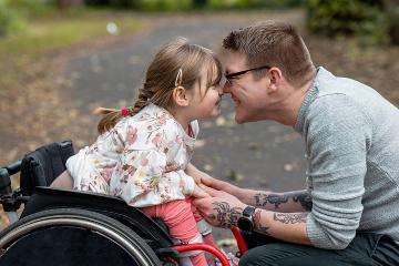 A close-up side view of a father and his young daughter who is a wheelchair user having a cute affectionate moment in a park