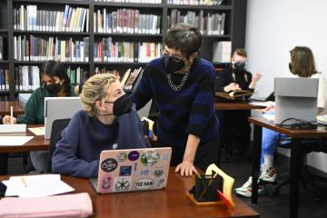 A student sits at a table with a laptop computer and speaks with an instructor standing next to her