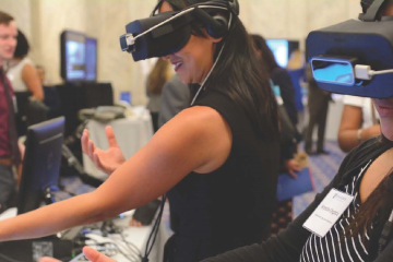 Photograph of a past Hopkins on the Hill event with two attendees having fun trying out virtual reality goggles. An illustration of the top of the Capitol building is in the bottom-right corner.