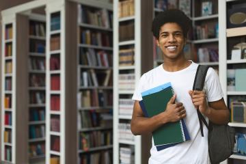 Young man holding notebooks and a backpack in an academic library with book stacks behind him.
