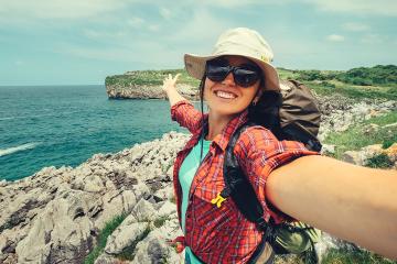 Young woman in hiking clothes standing on a rocky outcropping taking a selfie with a large body of water behind her 