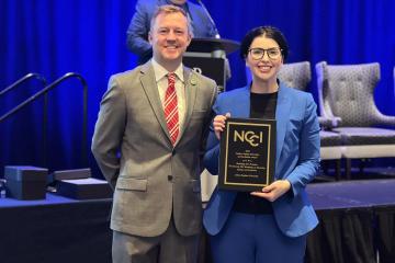 Two people pose for a photo, one holding an NCCI award plaque