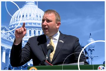 A man in a navy blue suit and yellow tie stands and speaks at a podium with the U.S. Capitol dome in the background