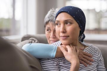 A female cancer patient with her head wrapped in a scarf is embraced from behind by an older woman