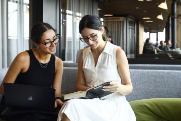 Two students sit on a plush bench while looking in a book