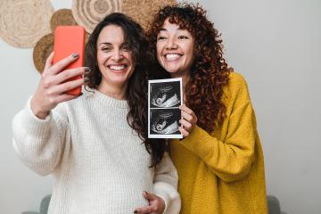 Two smiling women are Facetiming on a phone to show ultrasound images of their future child 
