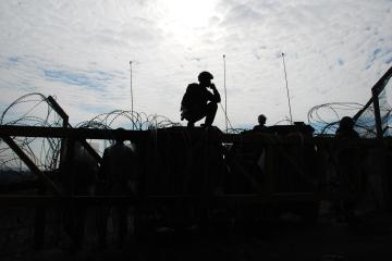 A soldier sits atop a barricade, silhouetted and surrounded by coils of barbed wire