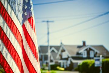 An American flag hanging from a house front porch on the 4th of July is in focus in the foreground, nice homes in the background and a clear blue sky