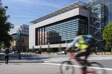 A large building with a glass facade is seen from across the street; a person on a bike rides past in the foreground