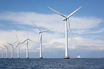 A row of giant wind turbines are shown in the ocean; a small sailboat is visible on the bottom right
