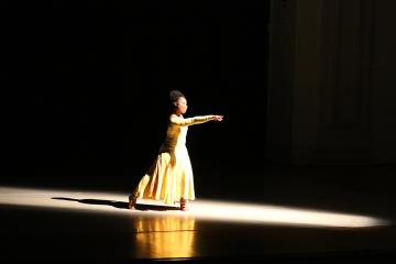 A solo dancer in a yellow dress in the middle of a spotlight on an otherwise dark stage