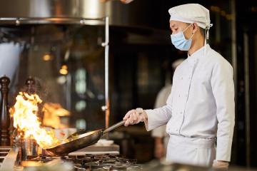 A male Asian chef wearing a surgical mask makes stir fry in a commercial kitchen
