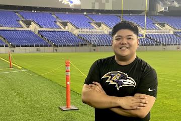 Mel Umahon stands on the sidelines of the football field at M&T Bank Stadium. Behind him, the Jumbotron reads 