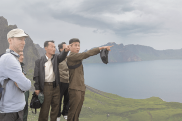 Four individuals stand on a green hillside near a large body of water; two of them are pointing at something our of frame to the right