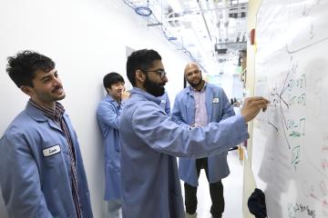 Researchers in blue lab coats gather around a whiteboard
