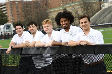 Five students in white t-shirts smile while leaning over a fence at Homewood Field.
