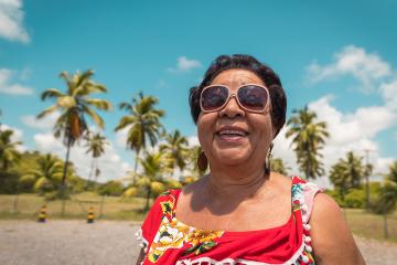 A smiling senior woman wearing sunglasses standing on a beach with palm trees behind her 