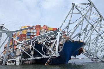 A large container ship pictured with the twisted steel remains of a collapsed bridge across its bow