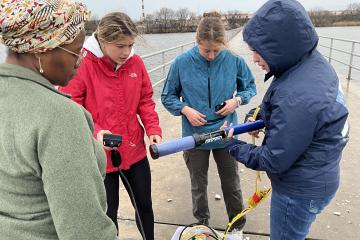 Four college students prepare scientific equipment on a dock next to the water.