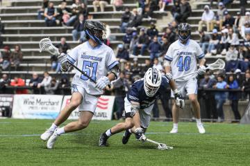 A Penn State men's lacrosse player tries to stop an opponent from Johns Hopkins. Behind them, another Johns Hopkins player watches.
