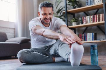 Man sitting on the floor at home stretches to touch his toes on one foot.