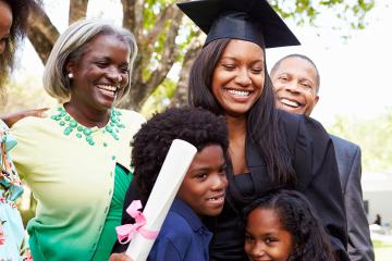 A woman wearing graduation cap and gown and holding a diploma hugs her children while her parents look on