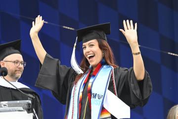 A student in a graduation cap and gown waves happily with both hands.