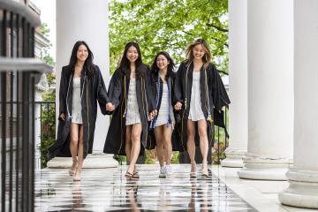 Four friends smile while walking hand in hand wearing black graduation robes