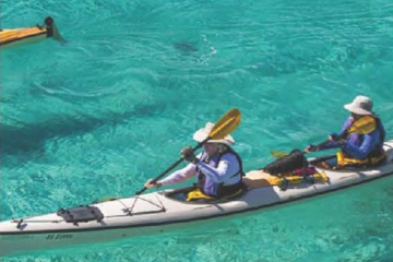 Photograph of two people in a kayak, paddling through turquoise-blue water