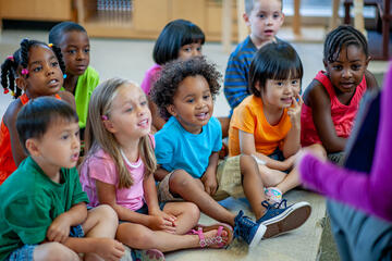 Toddlers sit on the floor while their teacher shows them a picture in a book.