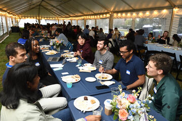 A group of people eat dinner at a long banquet table while talking amongst themselves.