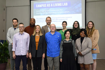 A group of 11 people stand in two rows while smiling for a group photo. Behind them, a projection on a screen reads "Campus as a Living Lab: Levering Our Campuses for Sustainability Research & Innovation."