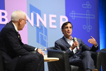 Ron Daniels speaks into a microphone while sitting next to Daniel M. Rubenstein. Both are wearing suits.